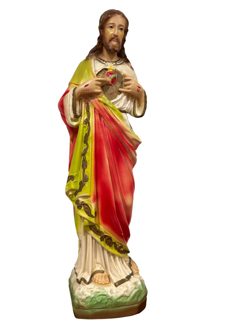 Photograph of a statue of the Sacred Heart of Jesus depicting him in a white robe with a gold-trimmed red sash. His Sacred Heart fills is shown on His chest and he is pointing to it.