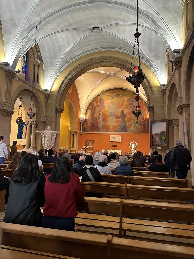 Photograph taken by Archbishop Etienne on his pilgrimage to France of believers kneeling praying in adoration in the Chapel of Apparitions. The chapel has wooden pews and yellow-gray stone walls. There is a white statue of the Jesus with his arms open in welcome on the left of the sanctuary. Above the sanctuary is a mural depicting Jesus revealing his Sacred Heart to St. Margaret Mary.
